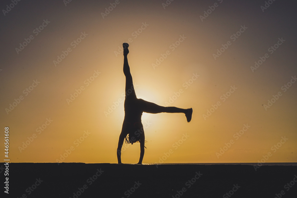 silhouette of a sportive athletic woman doing cartwheel on beach boardwalk during sunset 