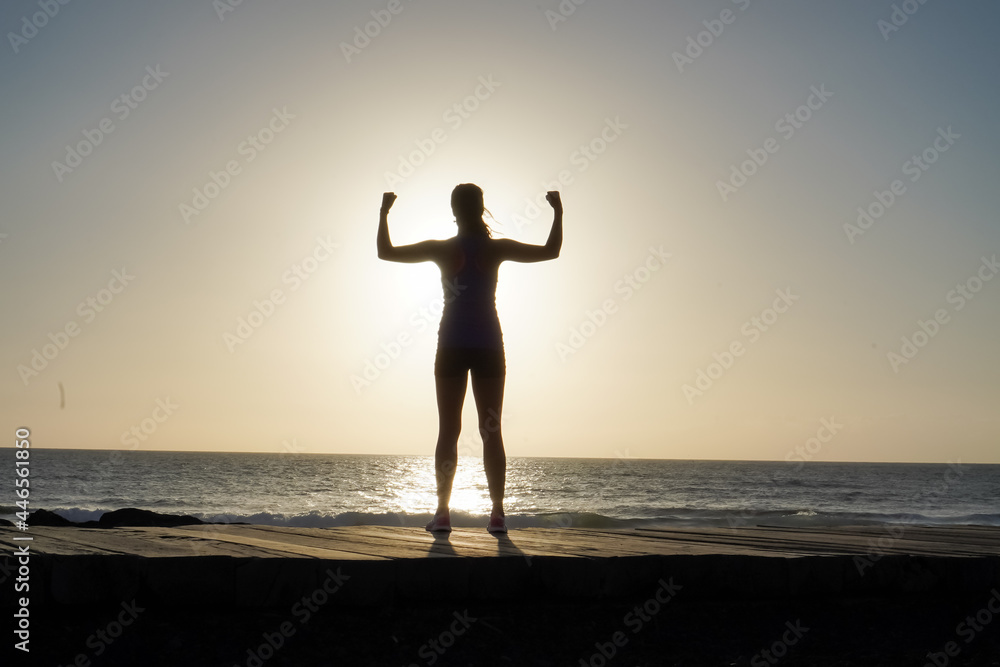 silhouette of a successful, strong  and sportive female doing champion pose with fists on beach boardwalk during sunset or sunrise 
