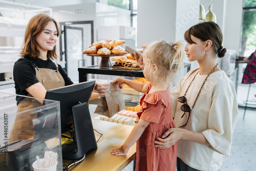 Fotografia Friendly cashier handing pastry goods in a bag to mom with a daughter