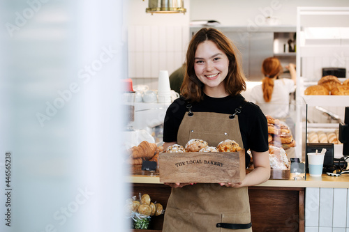 Obraz na plátně Friendly baker girl posing with a branded wooden box, filled with muffins