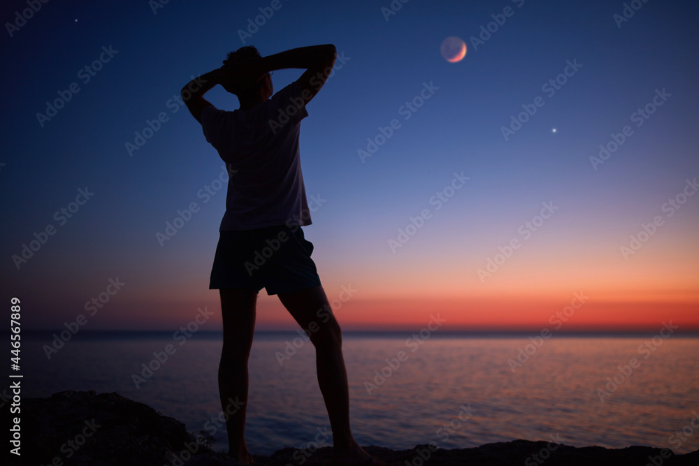 Silhouette of a man looking at the Moon and stars over sea ocean horizon.