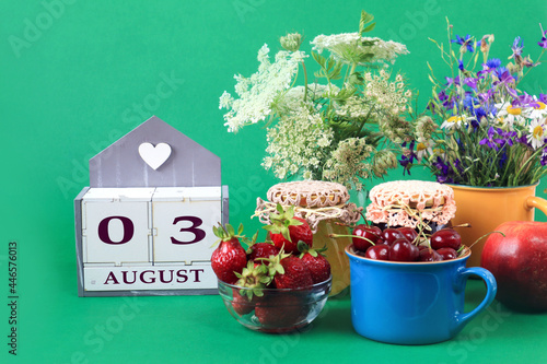 Calendar for August 3 : the name of the month of August in English, cubes with the numbers 0 and 3, bouquets of wild flowers, jars of jam, strawberries and cherries in cups, green background © MARYIA