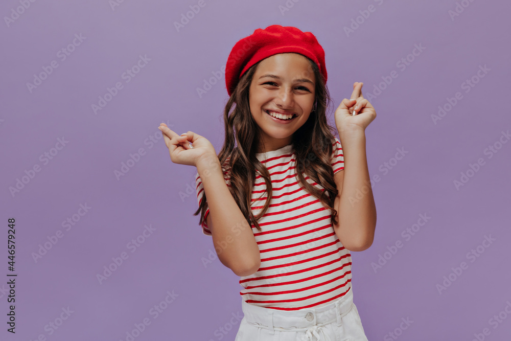 Pretty young girl crosses fingers on purple background. Attractive teenager in red beret and striped t-shirt smiles and looks into camera.