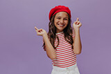 Pretty young girl crosses fingers on purple background. Attractive teenager in red beret and striped t-shirt smiles and looks into camera.