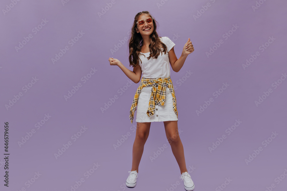 Active optimistic girl in white skirt and orange checkered shirt dances on purple background. Teenager smiles on isolated. Active optimistic girl in white skirt and orange checkered shirt dances on purple background. Teenager smiles on isolated.