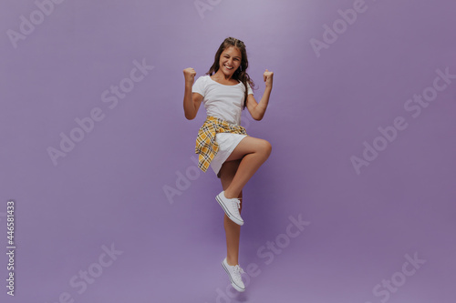 Excited tanned girl rejoicing on purple background. Teenager in white dress and checkered shirt jumps on isolated.