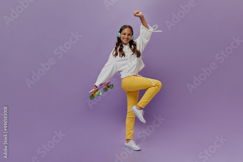 Active teenager moves on purple background and holds longboard. Young girl in yellow pants and white hoodie listens to music in headphones.