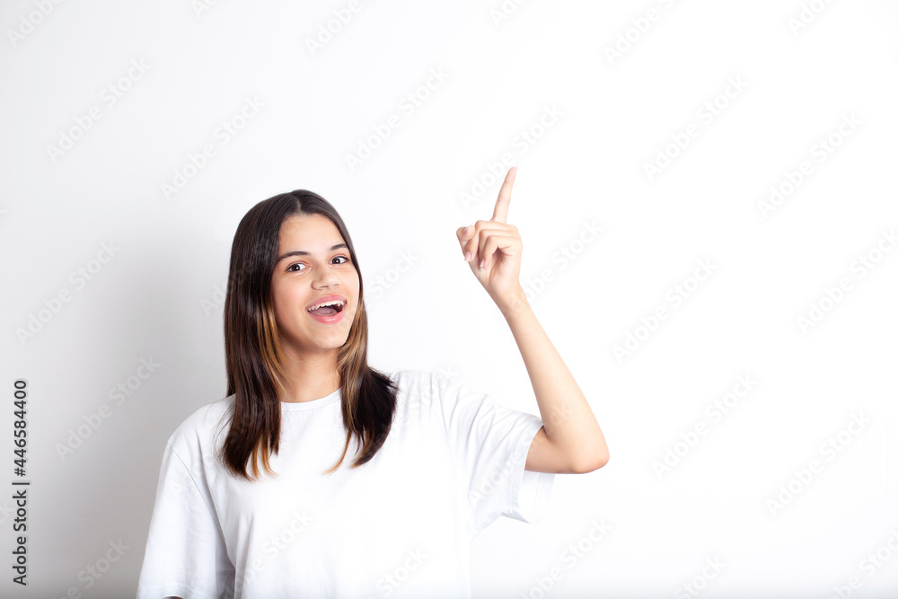A pretty teenage girl with dark hair with a smile points her finger up. A place for text. White background. Positive, joy, fun. A pretty teenage girl with dark hair with a smile points her finger up. A place for text. White background. Positive, joy, fun.