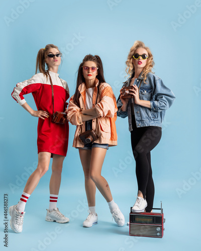Pretty young women in retro 90s fashion style, outfits posing isolated over blue studio background. Concept of eras comparison, beauty, fashion and youth. photo