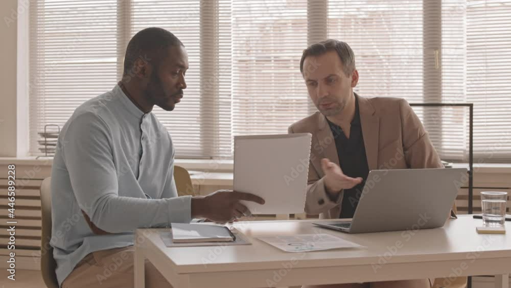 Medium long of middle-aged Caucasian man sitting at desk in office with his male African colleague, talking and working at portable computer and documents Medium long of middle-aged Caucasian man sitting at desk in office with his male African colleague, talking and working at portable computer and documents