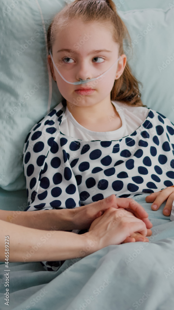 Hospitalized child wearing oxygen nasal tube resting in bed with medical oximeter on finger measuring heartbeat pulse. Worried mother discussing with kid during therapy consultation in hospital ward Hospitalized child wearing oxygen nasal tube resting in bed with medical oximeter on finger measuring heartbeat pulse. Worried mother discussing with kid during therapy consultation in hospital ward