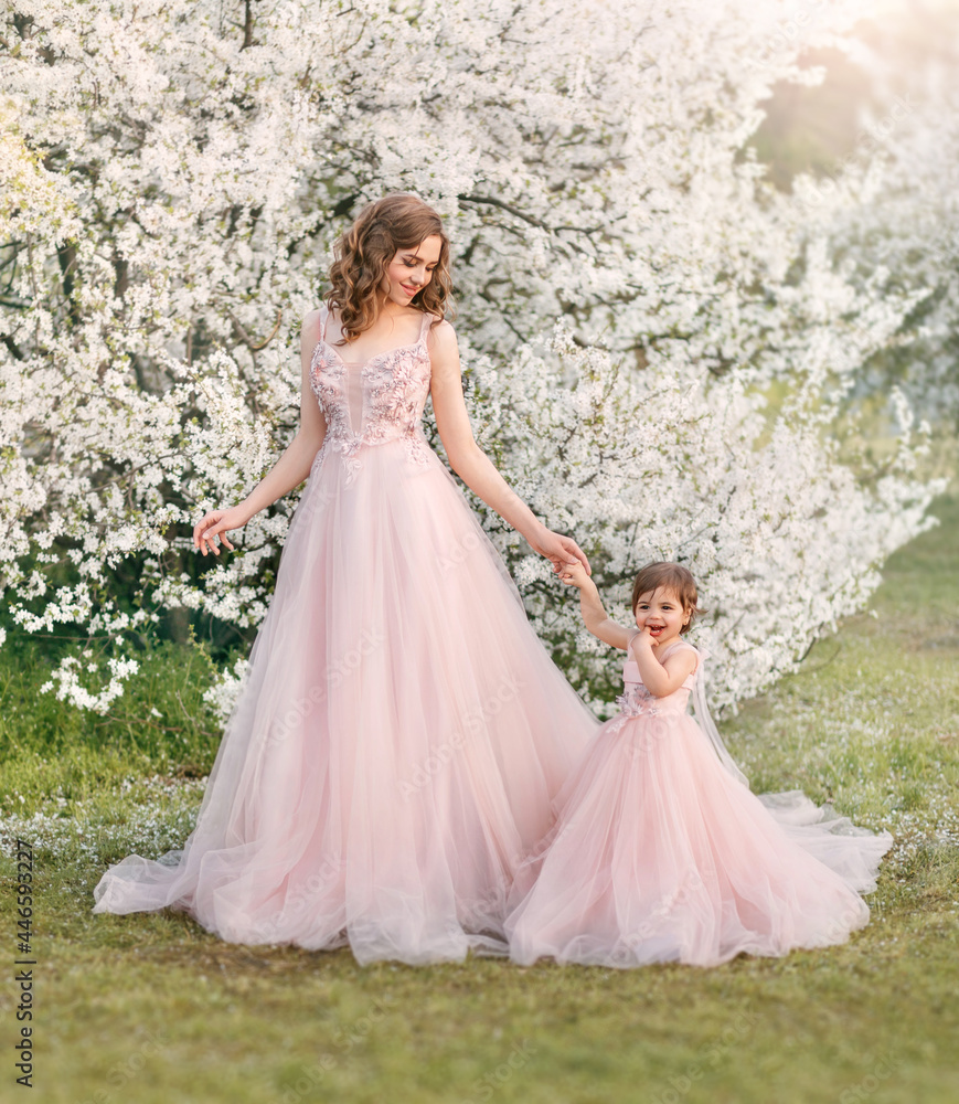 Beautiful fantasy woman gently holds hand little cheerful smiling girl. Happy family mom and daughter walk in blooming spring garden. Long wavy hair. Pink luxury vintage tulle fashion dress train