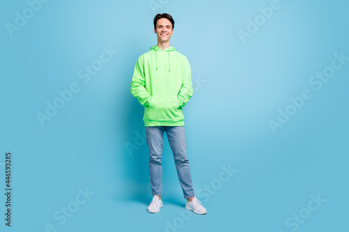 Photo of confident charming young guy dressed green hoodie arms pockets smiling isolated blue color background © deagreez
