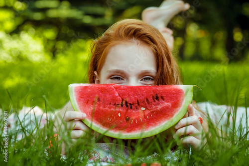 A beautiful girl with red hair eats a watermelon in nature on a warm summer day. Healthy lifestyle concept. Youth lifestyle. Funny photo.