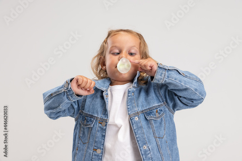 One little preshool Caucasian girl in denim clothes blowing bubble of gum isolated over white studio background. photo