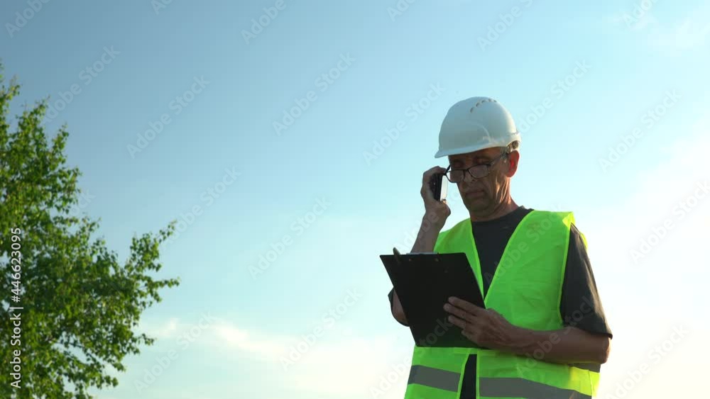 An elderly man wearing glasses and a work helmet makes notes against a clear blue sky An elderly man wearing glasses and a work helmet makes notes against a clear blue sky