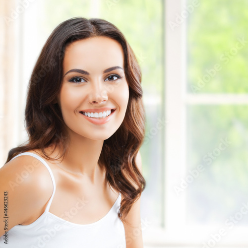 Portrait of happy smiling woman in white lingerie top sitting on bed, at bedroom, near window. Waking up attractive brunette girl at home room. Square.