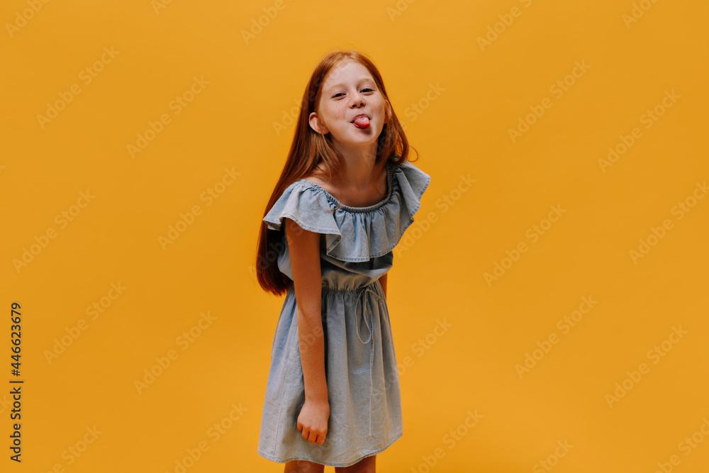 Cool little pretty girl in stylish blue dress shows tongue. Charming child makes funny face on orange isolated background. Cool little pretty girl in stylish blue dress shows tongue. Charming child makes funny face on orange isolated background.
