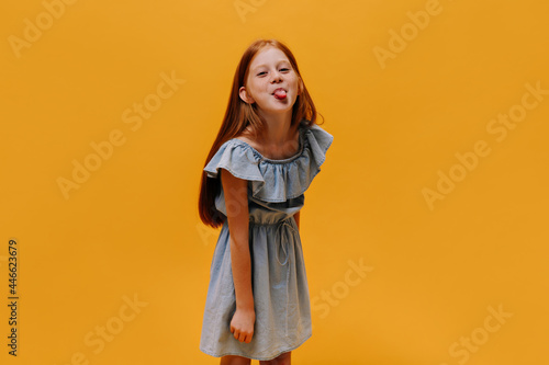 Cool little pretty girl in stylish blue dress shows tongue. Charming child makes funny face on orange isolated background.