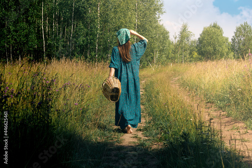 young woman in folk peasant clothes with a wicker basket walks along a field road