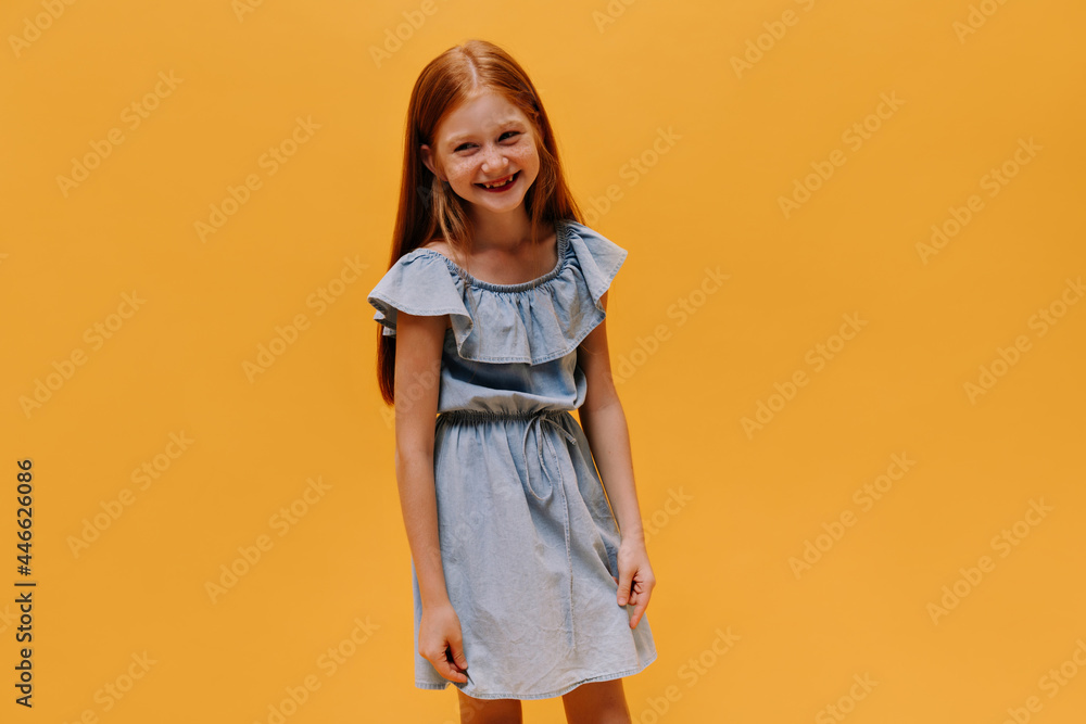 Portrait of happy excited long-haired redhead child in stylish denim dress smiling and posing on orange background. Portrait of happy excited long-haired redhead child in stylish denim dress smiling and posing on orange background.