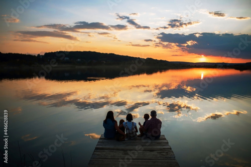 family of four people sitting on the shore on a wooden bridge of a large lake in summer and watching the beautiful sunset back view