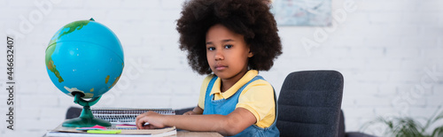 African american girl looking at camera near stationery and globe on table, banner