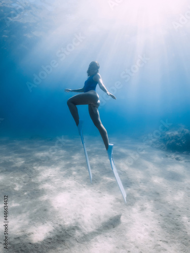 Female free diver with fins posing underwater in blue sea with sunlight.