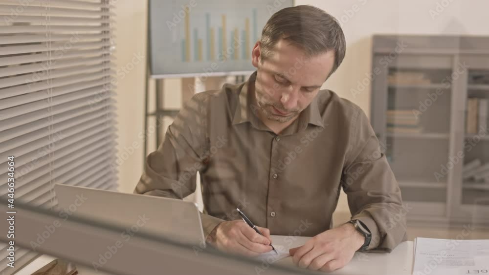 Waist-up of Caucasian male clerk sitting at desk in workplace, writing with pen on paper, using computer Waist-up of Caucasian male clerk sitting at desk in workplace, writing with pen on paper, using computer