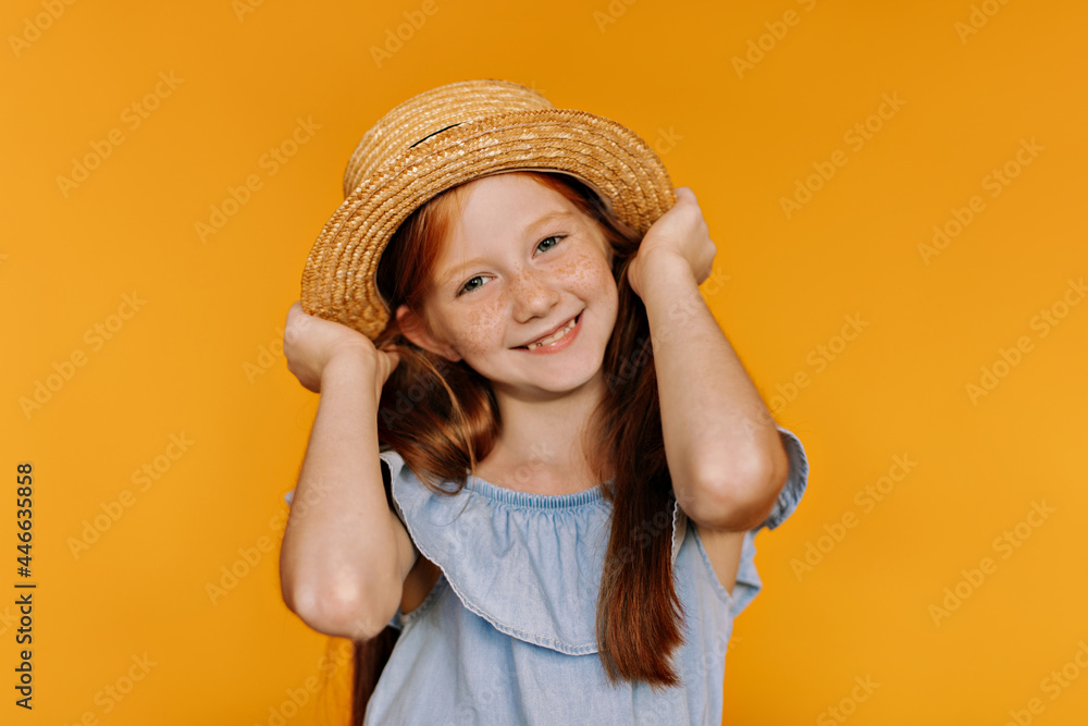 Beautiful long-haired redhead girl with freckles holds boater. Charming little child in blue blouse smiles on orange background. Beautiful long-haired redhead girl with freckles holds boater. Charming little child in blue blouse smiles on orange background.