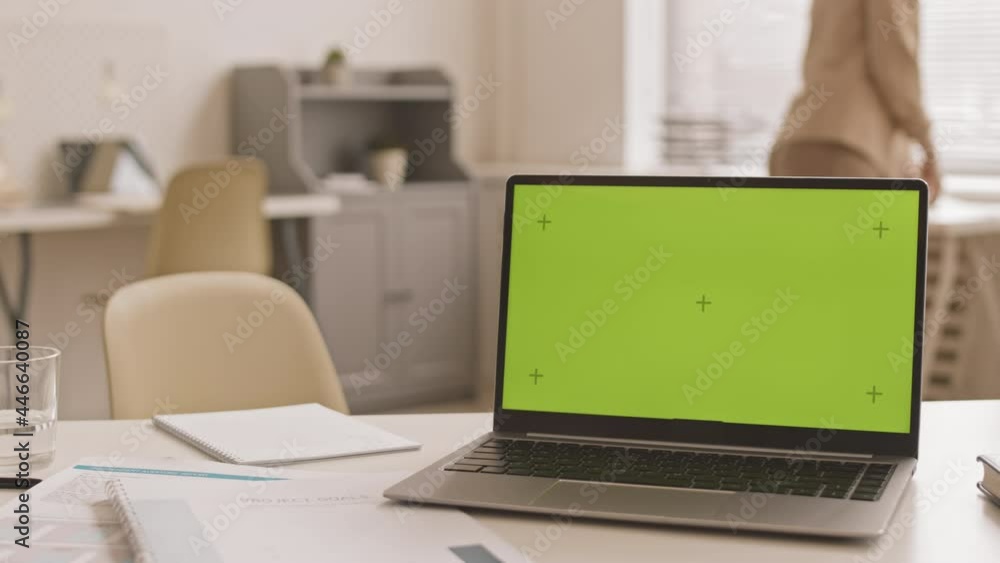 Tilting left of laptop computer with chroma key on screen on desk in office at daytime, unrecognizable woman with documents walking on background Tilting left of laptop computer with chroma key on screen on desk in office at daytime, unrecognizable woman with documents walking on background