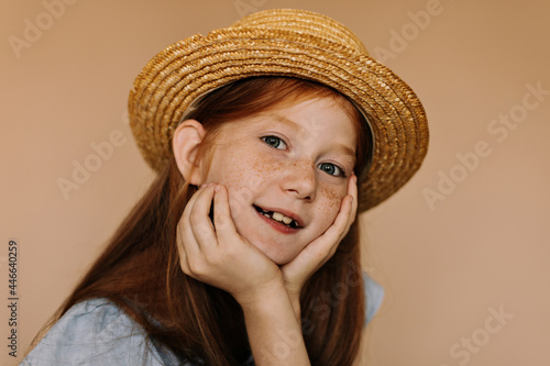 Portrait of lovely girl with freckles, grey eyes and red hair in modern hat and blue clothes looking into camera on beige backdrop..