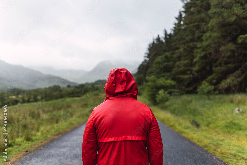 Man in red raincoat walking in an empty road surrounded by a forest during a cloudy day in the Highlands, Scotland
