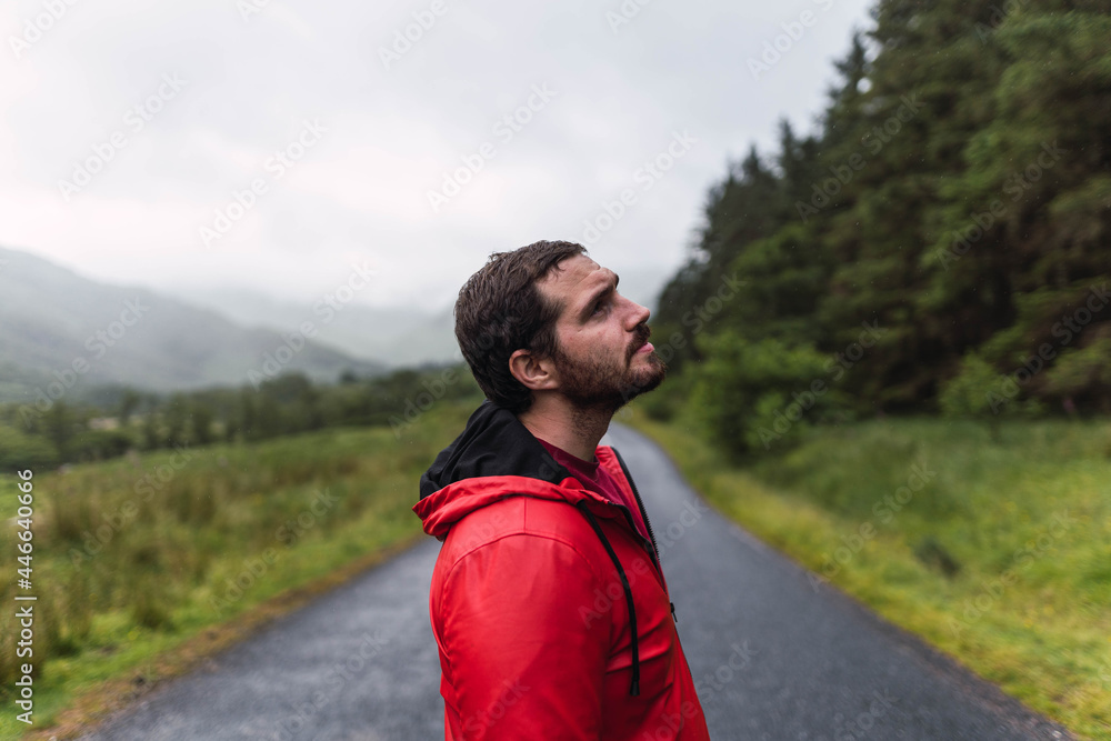 Man in red raincoat walking in an empty road surrounded by a forest during a cloudy day in the Highlands, Scotland