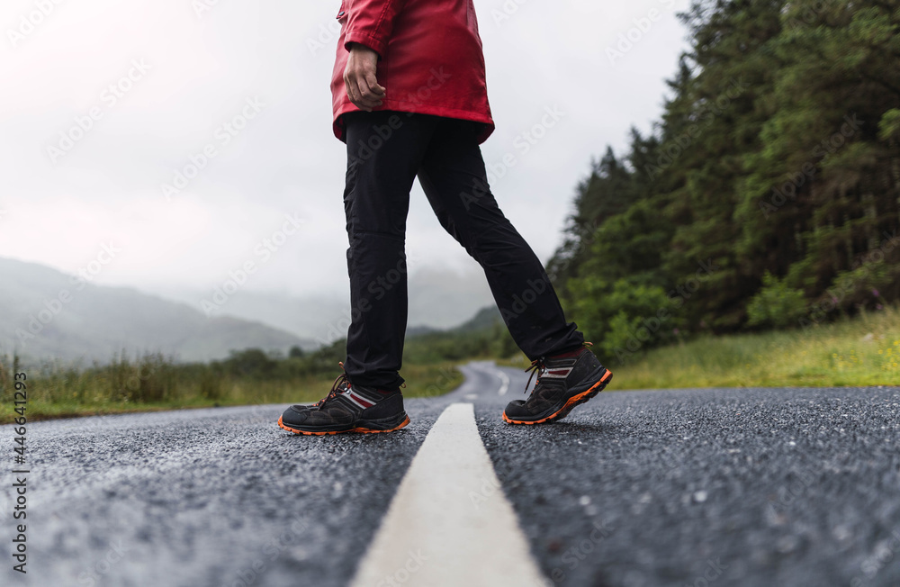 Traveler walking into the horizon on the lonely road in Scotland enjoying the scenery. He is wearing a red raincoat and surrounded by green trees and nature. Enjoying freedom