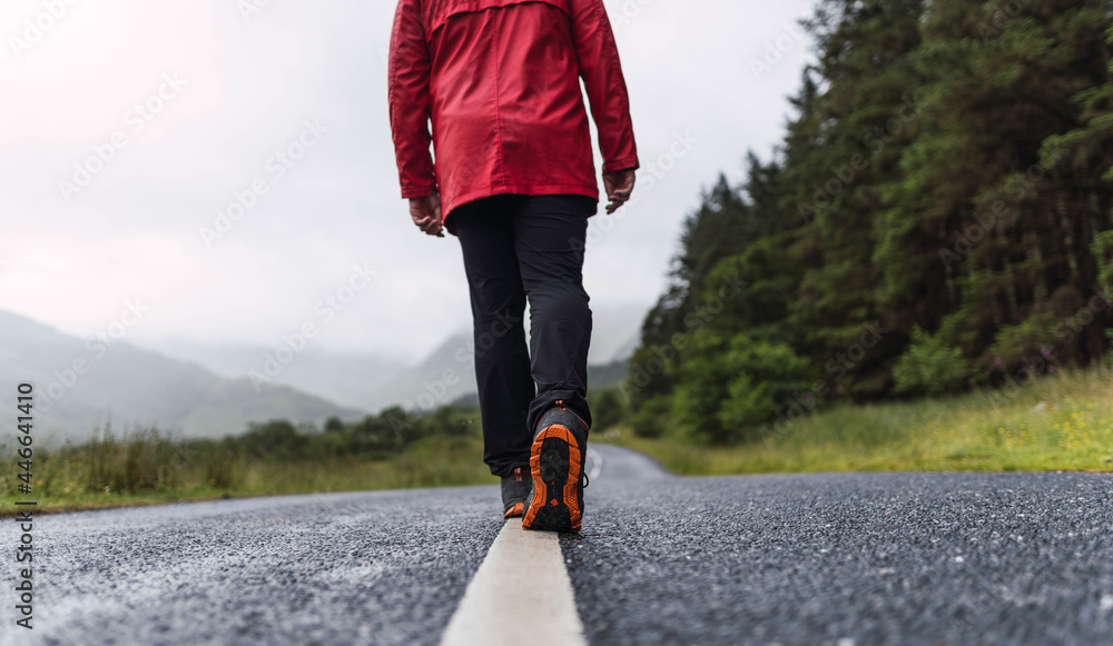 Traveler walking into the horizon on the lonely road in Scotland enjoying the scenery. He is wearing a red raincoat and surrounded by green trees and nature. Enjoying freedom