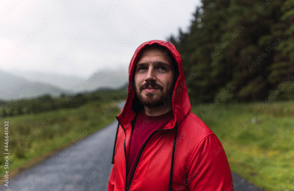 Man in red raincoat walking in an empty road surrounded by a forest during a cloudy day in the Highlands, Scotland