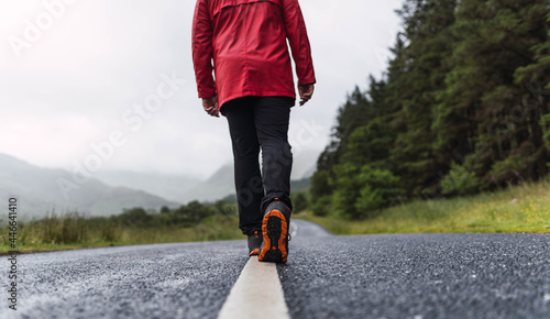 Obraz na płótnie Traveler walking into the horizon on the lonely road in Scotland enjoying the scenery. He is wearing a red raincoat and surrounded by green trees and nature. Enjoying freedom
w salonie