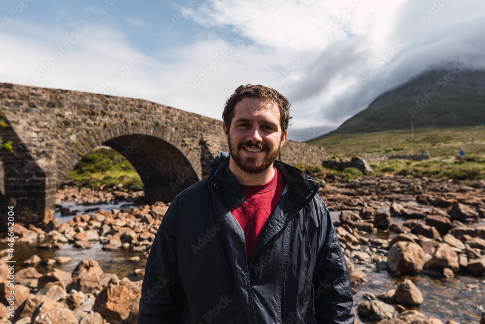 Young man sitting on the rock bridge while enjoying Scotland hill view at the Highlands, in Scotland. He is wearing hiking clothes. Sitting on an old vintage bridge in Sligachan, Isle of Skye