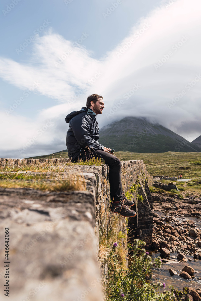 Young man sitting on the rock bridge while enjoying Scotland hill view at the Highlands, in Scotland. He is wearing hiking clothes. Sitting on an old vintage bridge in Sligachan, Isle of Skye