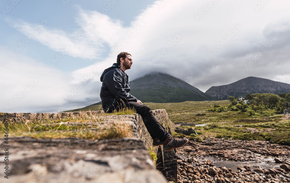 Young man sitting on the rock bridge while enjoying Scotland hill view at the Highlands, in Scotland. He is wearing hiking clothes. Sitting on an old vintage bridge in Sligachan, Isle of Skye