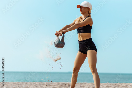 Strong young woman, training the muscles of the arms and legs with the help of kettlebells, woman in sportswear, training on the beach