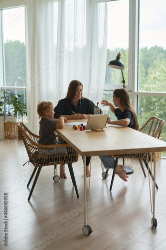  mom and her children girls are sitting at the table and drawing with paints using an online lesson on a tablet           
