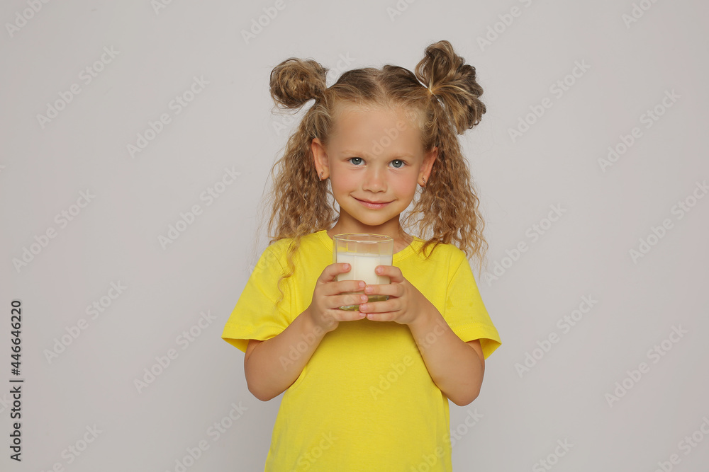 a beautiful blonde girl with ponytails in a yellow T-shirt holds a glass glass with milk on a white background