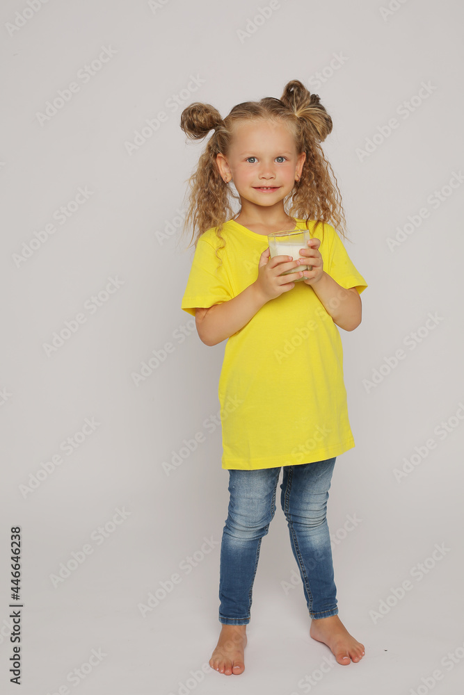 a beautiful blonde girl with ponytails in a yellow T-shirt holds a glass glass with milk on a white background a beautiful blonde girl with ponytails in a yellow T-shirt holds a glass glass with milk on a white background