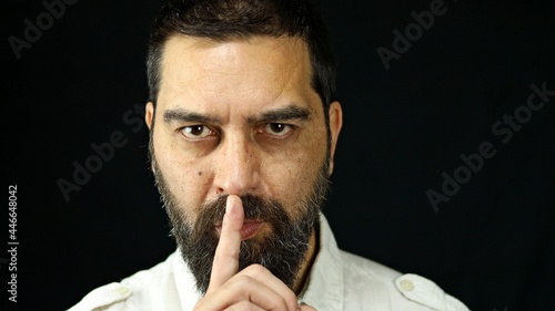 Man with beard on black background holding index finger in front of mouth making silence sign. Silence concept