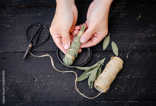 Person making white sage Salvia apiana smudge stick at home with homegrown sage leaves. Cotton string and vintage scissors for decoration on black wood background. Flat lay view. photo