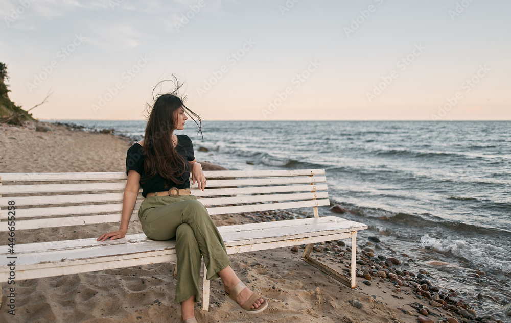 Beautiful brunette woman looking far into the sea horizon. Girls sits on white bench on the beach. Concept of work-life balance and wellbeing. Beautiful brunette woman looking far into the sea horizon. Girls sits on white bench on the beach. Concept of work-life balance and wellbeing.