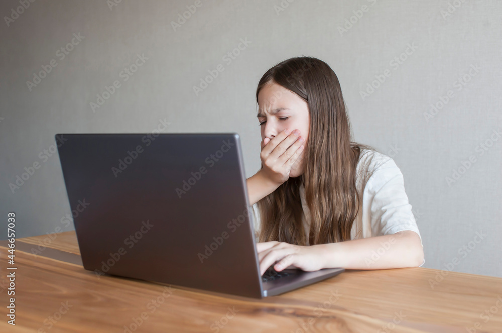 Little schoolgirl girl is tired and yawns using laptop on the table at home. Online education of the student. Fatigue. Back to school Little schoolgirl girl is tired and yawns using laptop on the table at home. Online education of the student. Fatigue. Back to school