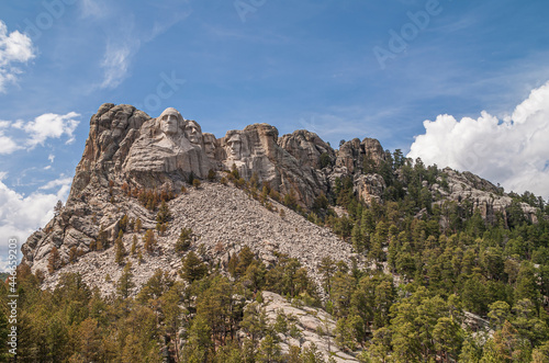 Black Hills, Keystone, SD, USA - May 31, 2008: Mount Rushmore sculpture far up the mountain slope under blue cloudscape, Green forest on gray granite.